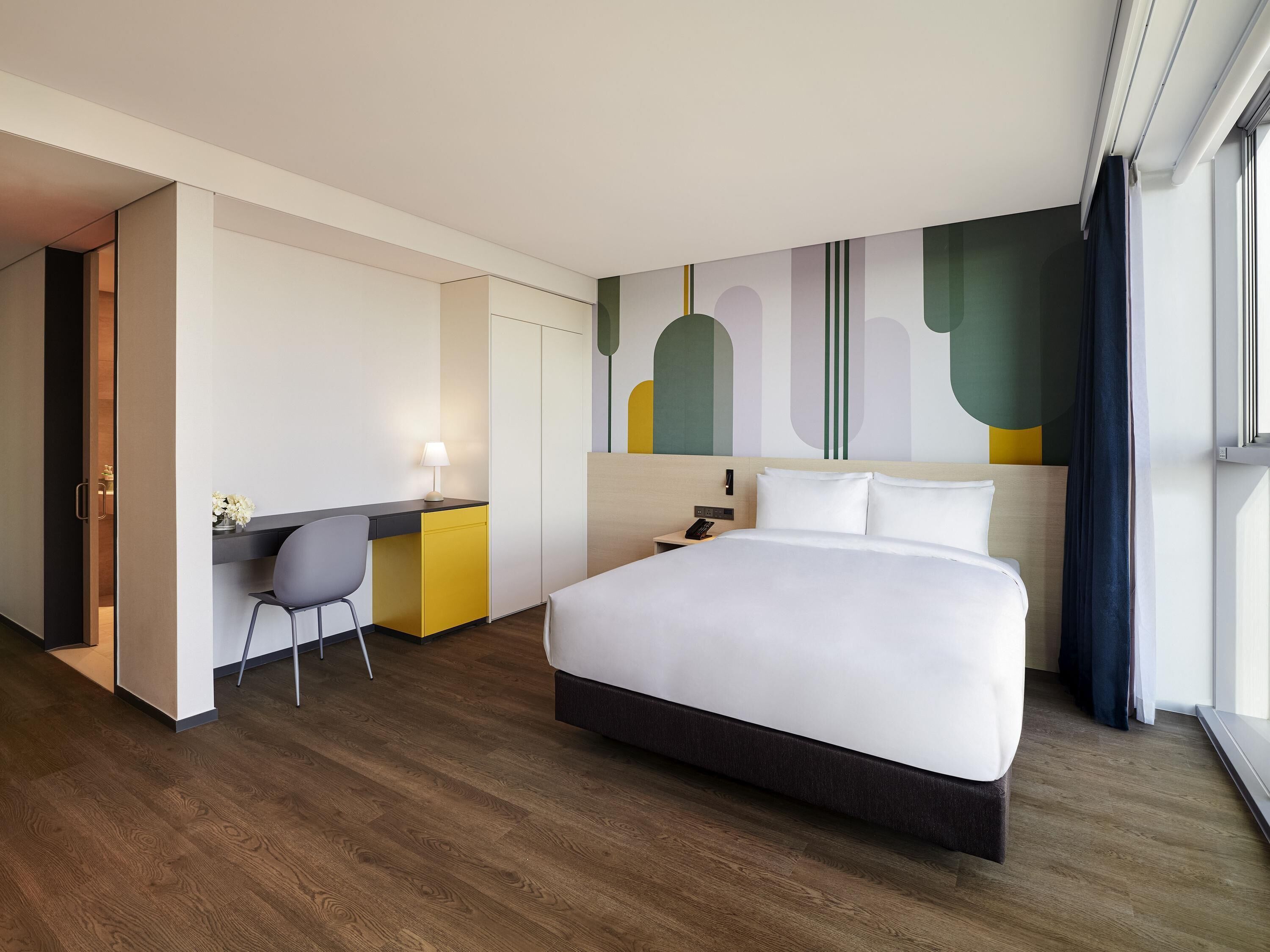 A hotel room with a large bed, a desk with a chair, a nightstand, and a wall with abstract art.