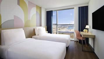 Une chambre d’hôtel avec deux lits, un bureau avec une lampe, une chaise et une grande fenêtre donnant sur l’aéroport.