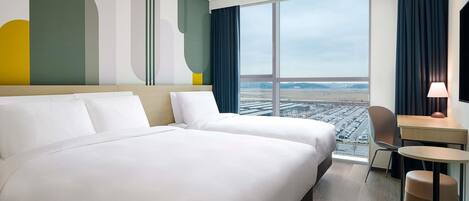 Une chambre d’hôtel avec deux lits, un bureau, une chaise, une lampe et une grande fenêtre donnant sur un aéroport.