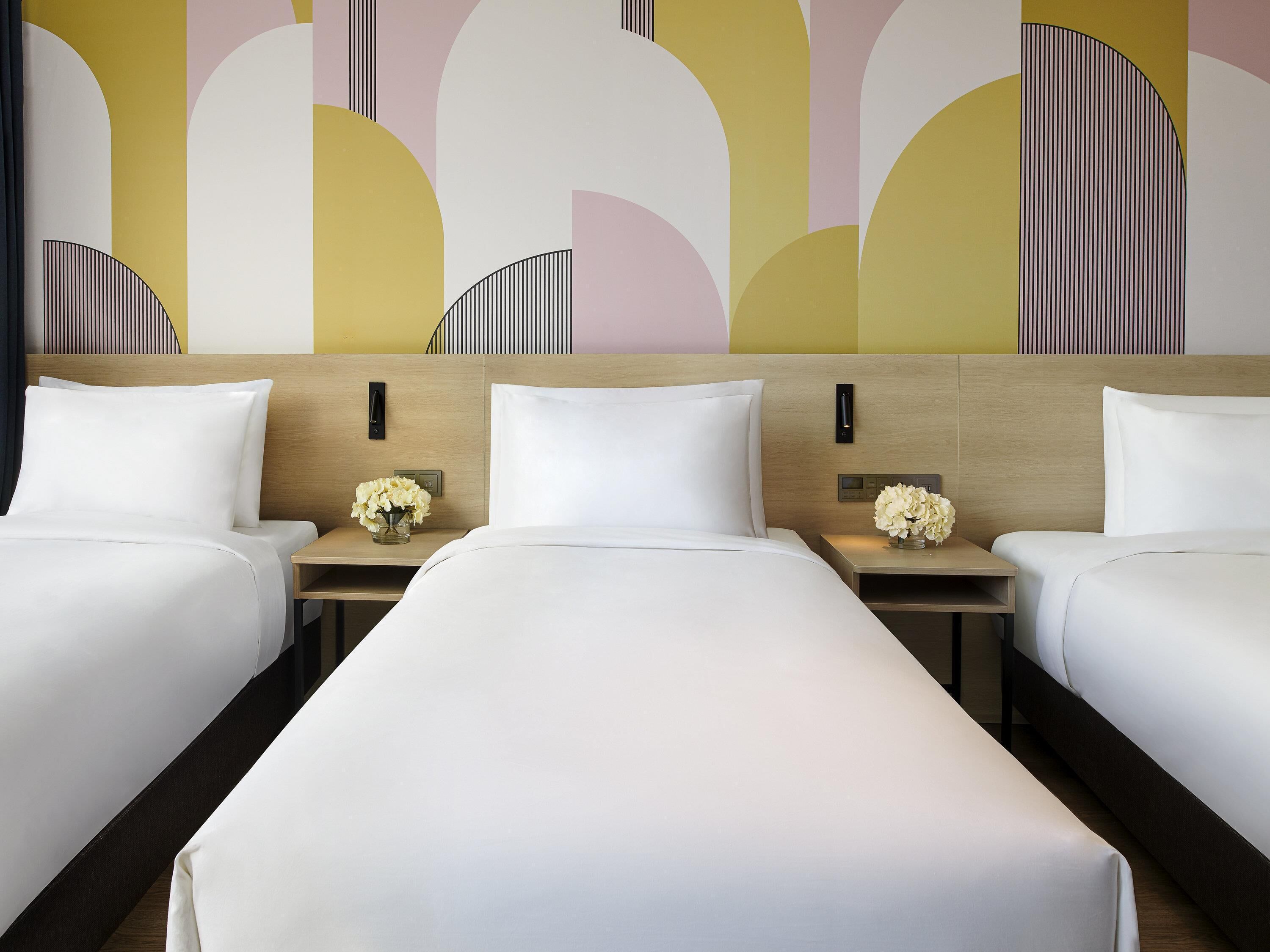A hotel room with three beds, a wooden headboard, bedside tables with lamps, and a decorative wall.