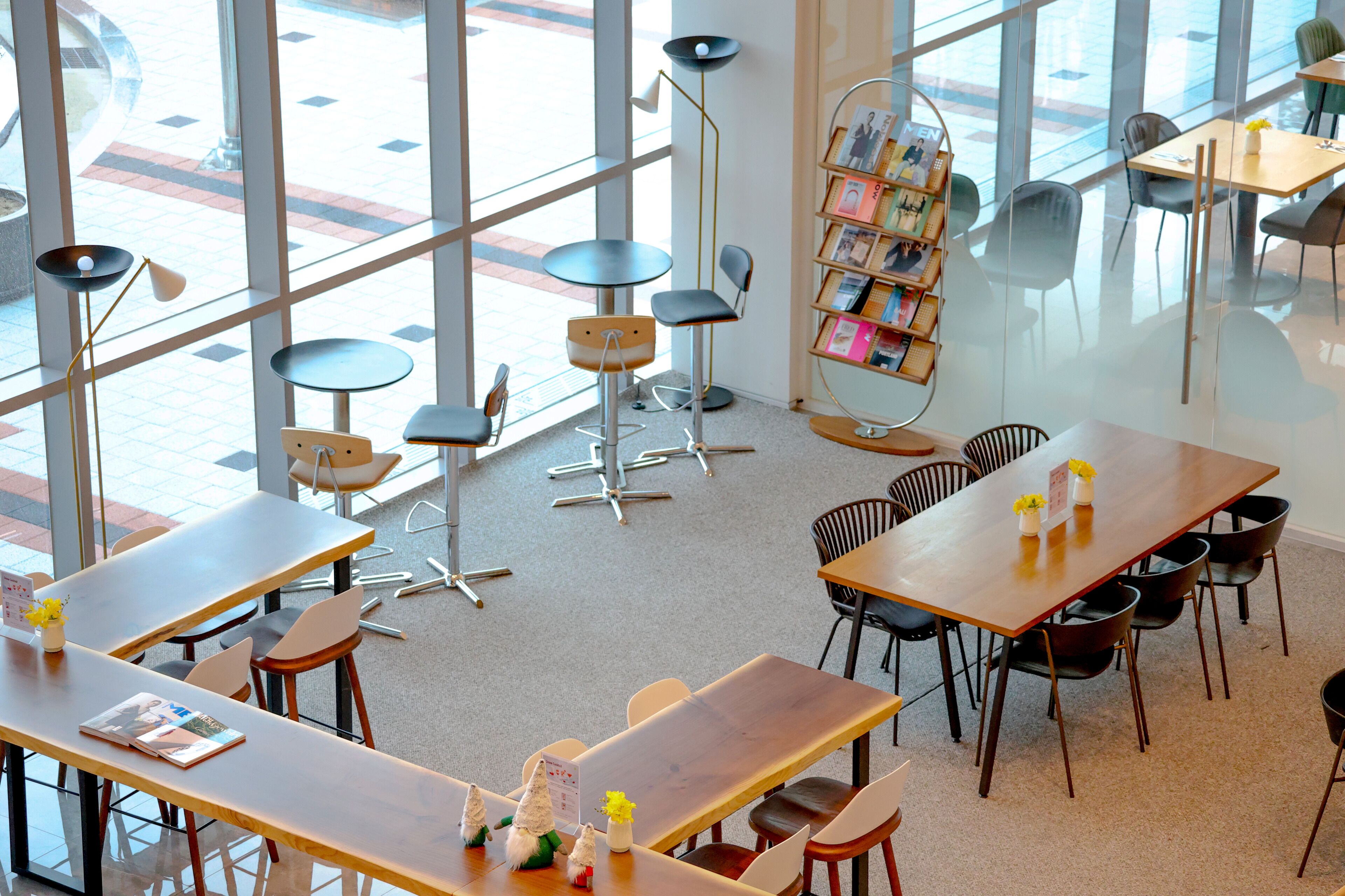 Un café moderne doté de nombreuses tables et chaises, d’une bibliothèque avec des magazines et de grandes fenêtres donnant sur la rue.