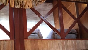Ein Zimmer mit Holzbalkendecke, einem Bett mit blau-weißem Kopfkissen und einer Strohmatte, die von der Decke hängt.