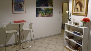 Una habitación con un escritorio blanco, dos sillas blancas, una estantería con libros y objetos decorativos, y tres obras de arte enmarcadas en la pared.