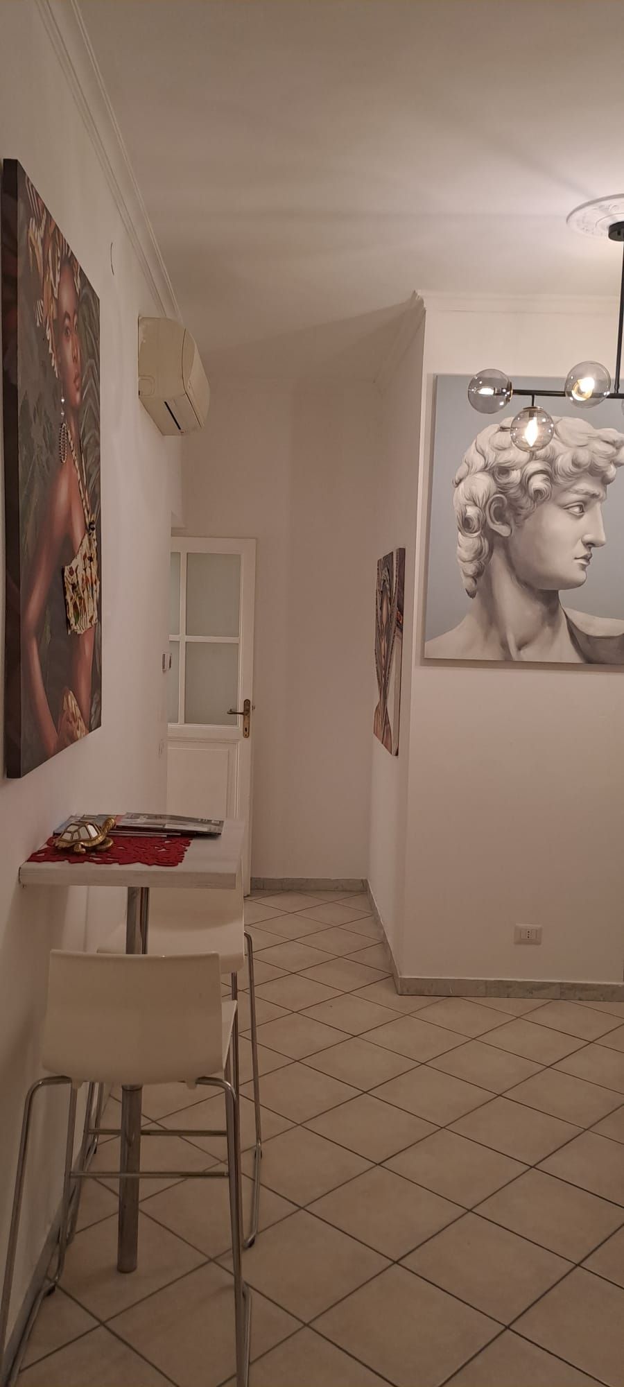 Une pièce avec un sol carrelé, une table blanche avec une chaise, une porte et deux œuvres d’art encadrées accrochées aux murs.