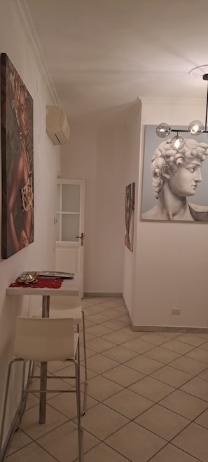 A room with a tiled floor, a white table with a chair, a door, and two framed artworks on the walls. - Apartment (Roma)
