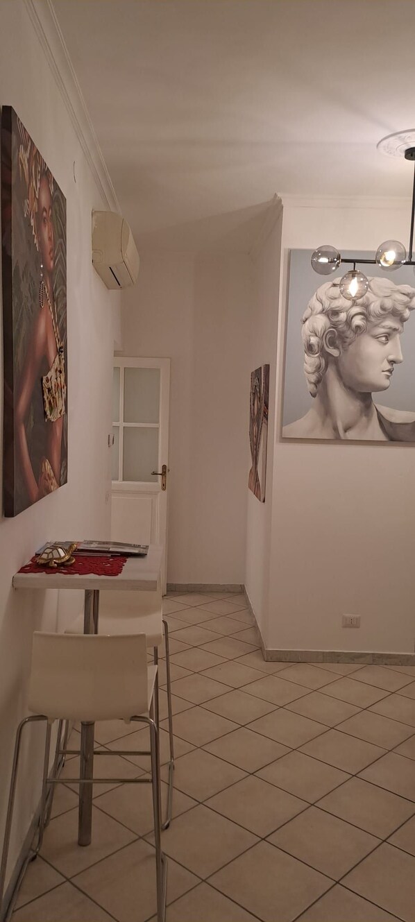 Une pièce avec un sol carrelé, une table blanche avec une chaise, une porte et deux œuvres d’art encadrées accrochées aux murs.