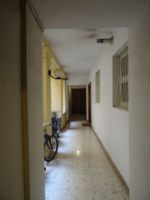 A narrow hallway with a bicycle parked on the left, a door at the end, and a barred window on the right.
