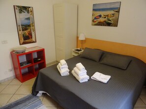 Una cama bien hecha con toallas, un estante rojo con libros, un cuadro de una playa y una lámpara en una mesita de noche.