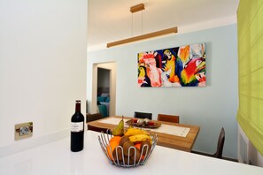 Un coin repas avec une table dressée pour deux personnes, un saladier de fruits et une bouteille de vin. Un tableau coloré est accroché au mur, au-dessus de la table.