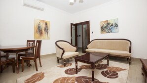 A living room with a sofa, two armchairs, a coffee table, and a dining table with chairs. There are two paintings on the walls and a door leading to another room.