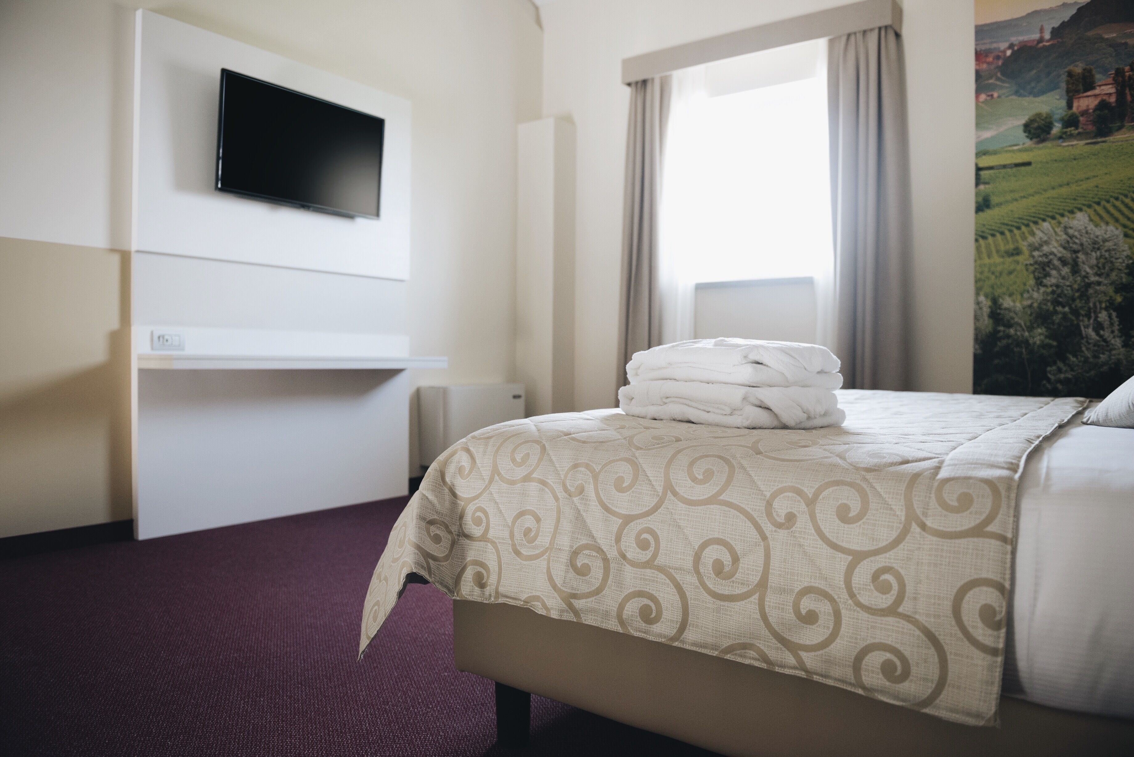 A hotel room with a bed, a flat-screen TV mounted on the wall, a window with curtains, and a painting on the wall.