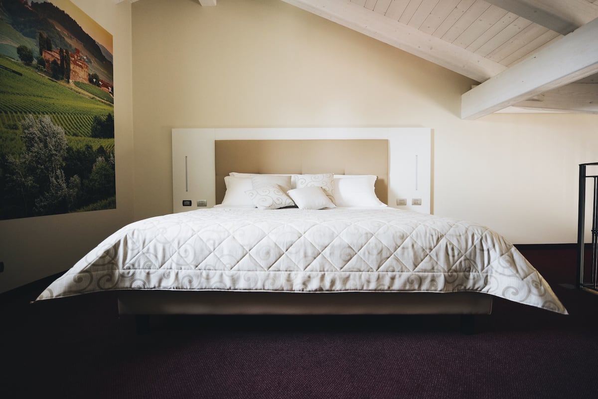 A neatly made bed with a quilted white duvet and matching pillows, a headboard with wall-mounted lights, and a large landscape painting on the wall.