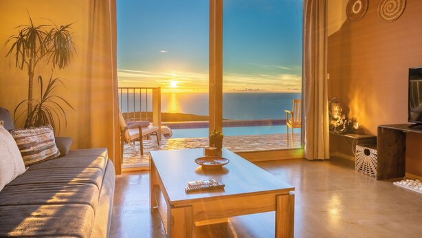 A living room with a sofa, a coffee table, and a TV stand. There is a large window with a view of the sunset and the ocean. - Villa Dioni-Ocean view from every room, spacious and very clean. (Magganos)