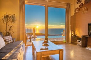 Un salon comprenant un canapé, une table basse et un meuble pour la télévision. Une grande fenêtre offre une vue sur le coucher du soleil et l’océan.