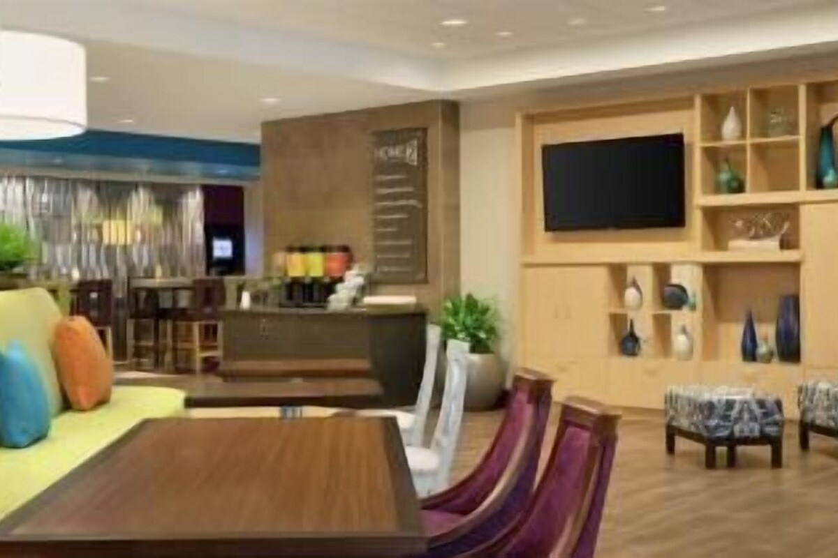 A modern hotel lobby with a sofa, dining table, chairs, a TV, and a bar area.