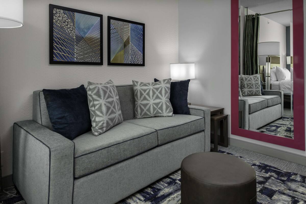Une chambre d’hôtel moderne avec un canapé gris, des coussins à motifs, un pouf rond et deux tableaux abstraits accrochés au mur.