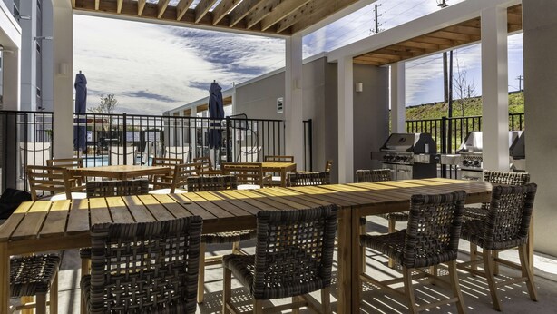 An outdoor dining area with wooden tables and wicker chairs, a barbecue grill, and a view of a hill. - Home2 Suites by Hilton Covington (Covington)