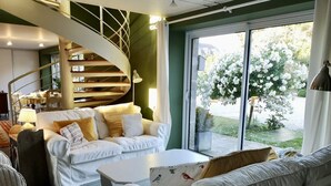 Een moderne woonkamer met een witte bank, een grijze bank en een witte salontafel. Achtergrond is een spiltrap en een groot raam met uitzicht op een tuin.