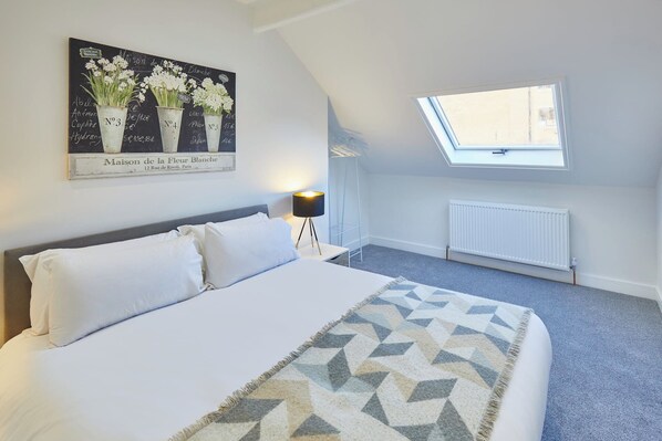 A bedroom with a bed, a nightstand, a lamp, a window, and a wall-mounted painting.