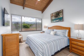 Un dormitorio con un ventanal grande, una cama con ropa de cama a rayas, cabecero de madera, una mesita de noche con una lámpara y una mesita auxiliar de mimbre.
