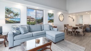 A modern living room with a sectional sofa, dining table, and chairs. There are paintings on the walls and a large window with blinds.