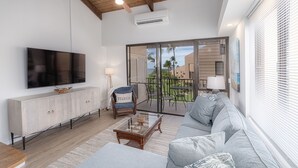A modern living room with a flat-screen TV, a sofa, a coffee table, and a chair. There is a balcony with a view of palm trees and buildings.
