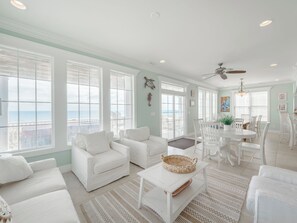 A spacious living room with white furniture, large windows with ocean views, and a dining area with white chairs and a table.