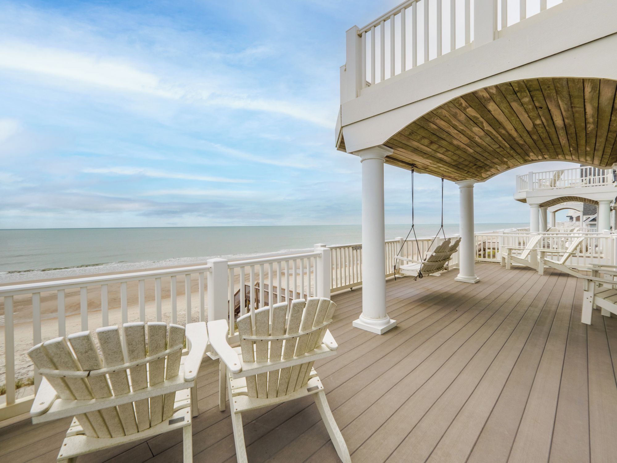 A wooden deck with white railings and chairs overlooking a beach.