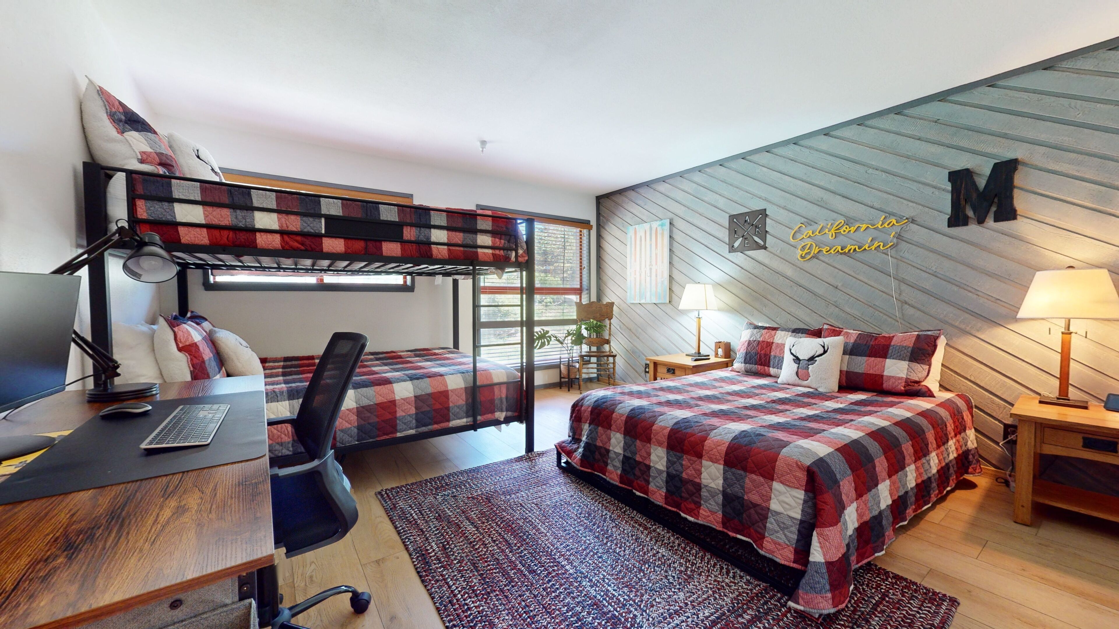 A dorm-style room with bunk beds, a desk with a computer, a chair, a bed with a plaid bedspread, a nightstand with a lamp, and a wall with a 'M' sign.
