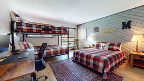 A dorm-style room with bunk beds, a desk with a computer, a chair, a bed with a plaid bedspread, a nightstand with a lamp, and a wall with a 'M' sign. - Aspen Creek #115 2 Bedroom Condo by RedAwning (Mammoth Lakes)