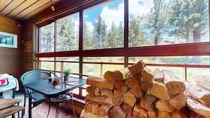 A cozy cabin interior with a large window, a table with chairs, and a stack of firewood.