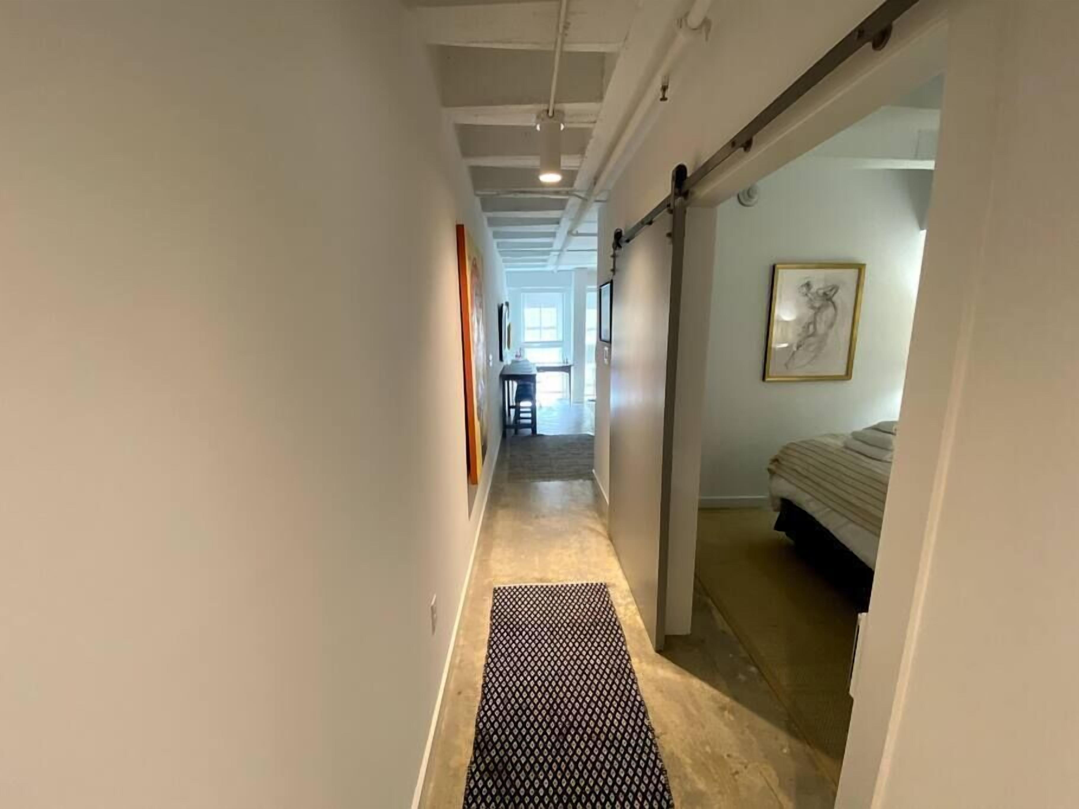 A narrow hallway with a black patterned mat, white walls, and a bed with a headboard visible in the adjacent room.