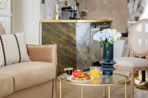 Un salon moderne avec un canapé beige, une table basse en verre sur laquelle sont disposés des éléments pour le petit-déjeuner, et un vase décoratif rempli de fleurs.