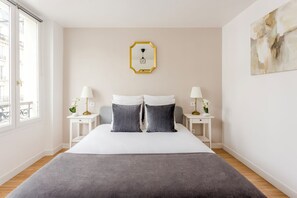 Ein modernes Hotelzimmer mit einem großen Bett, zwei Nachttischen, einer Wandleuchte und Blick auf die Stadt durch das Fenster.