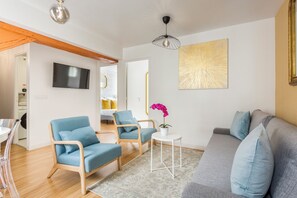 Ein modernes Wohnzimmer mit einem grauen Sofa, zwei blauen Sesseln, einem kleinen weißen Tisch mit Topfpflanze und einem an der Wand befestigten Fernseher.