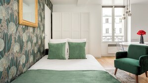 Une chambre à coucher avec un lit, des coussins verts, un fauteuil vert, un miroir et une fenêtre donnant sur des immeubles.