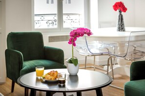 A round table with a green chair, a white vase with pink flowers, a glass of orange juice, a plate with croissants, and a glass of coffee.