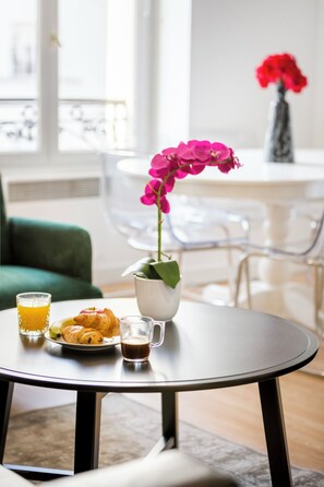 A dining table with a plate of croissants, a glass of orange juice, and a cup of coffee. There is a vase with a pink orchid on the table. - Merveil - Family Suite - Marais - Martin VI (Paris)