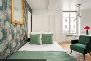 A bedroom with a bed, green pillows, a green chair, a mirror, and a window with a view. - Merveil - Family Suite - Marais - Martin VI (Paris)