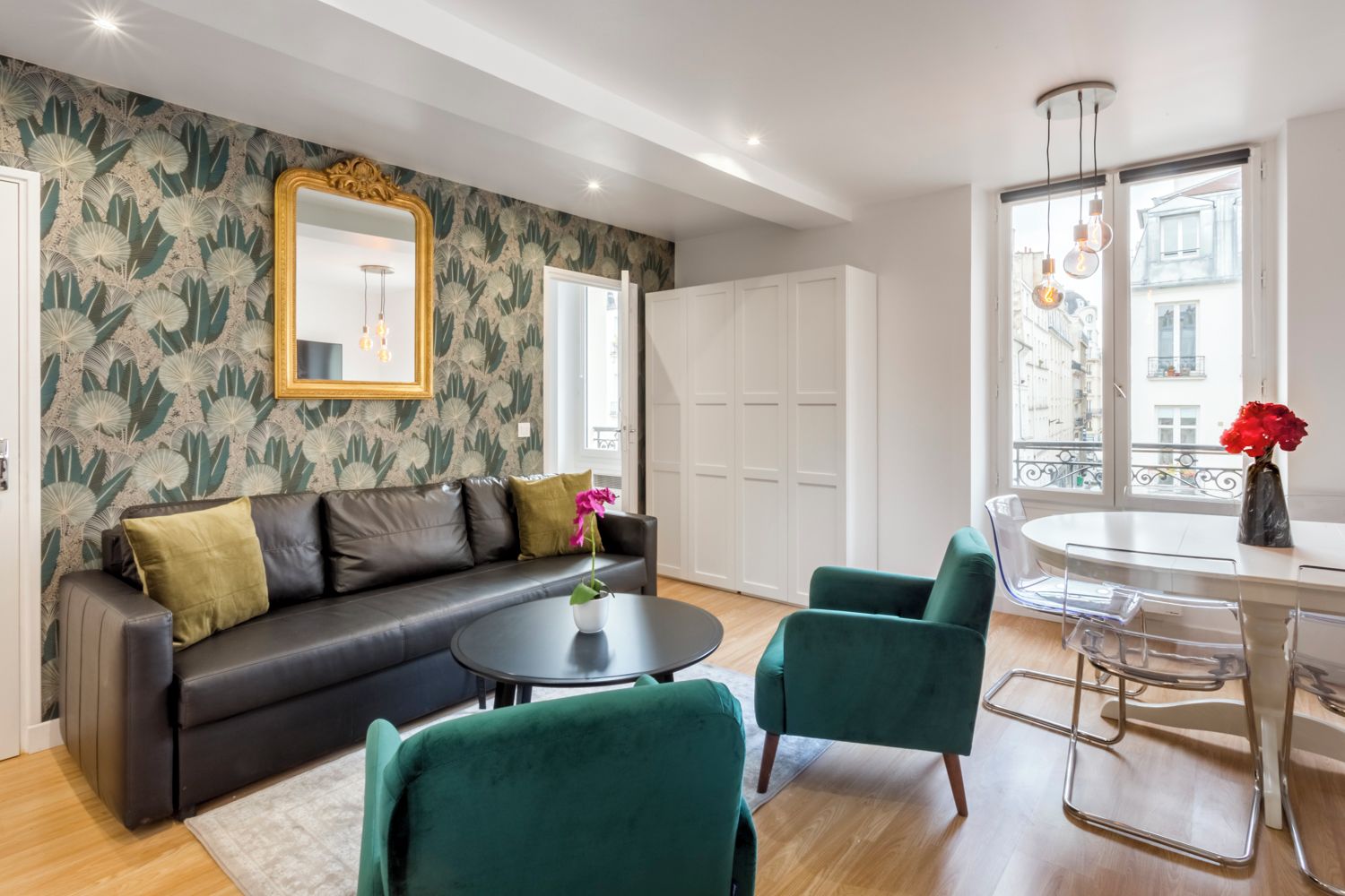 A modern living room with a black leather sofa, green armchairs, a round coffee table, and a dining table with chairs. There is a large mirror on the wall, a window with a view of buildings, and a vase with flowers on the dining table.