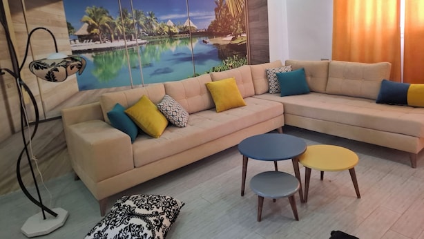 Un salón con un sofá seccional color crema, dos mesas redondas y un mural grande con una escena tropical.