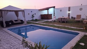 Una piscina con plataforma de baldosas, un área de estar techada y un pequeño jardín con plantas en macetas.