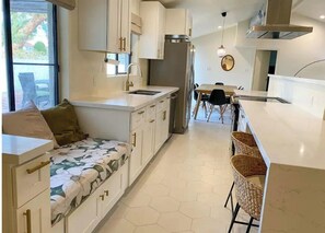 Una cocina moderna con armarios blancos, un refrigerador de acero inoxidable, un comedor con mesa y sillas, y un ventanal que proporciona luz natural.