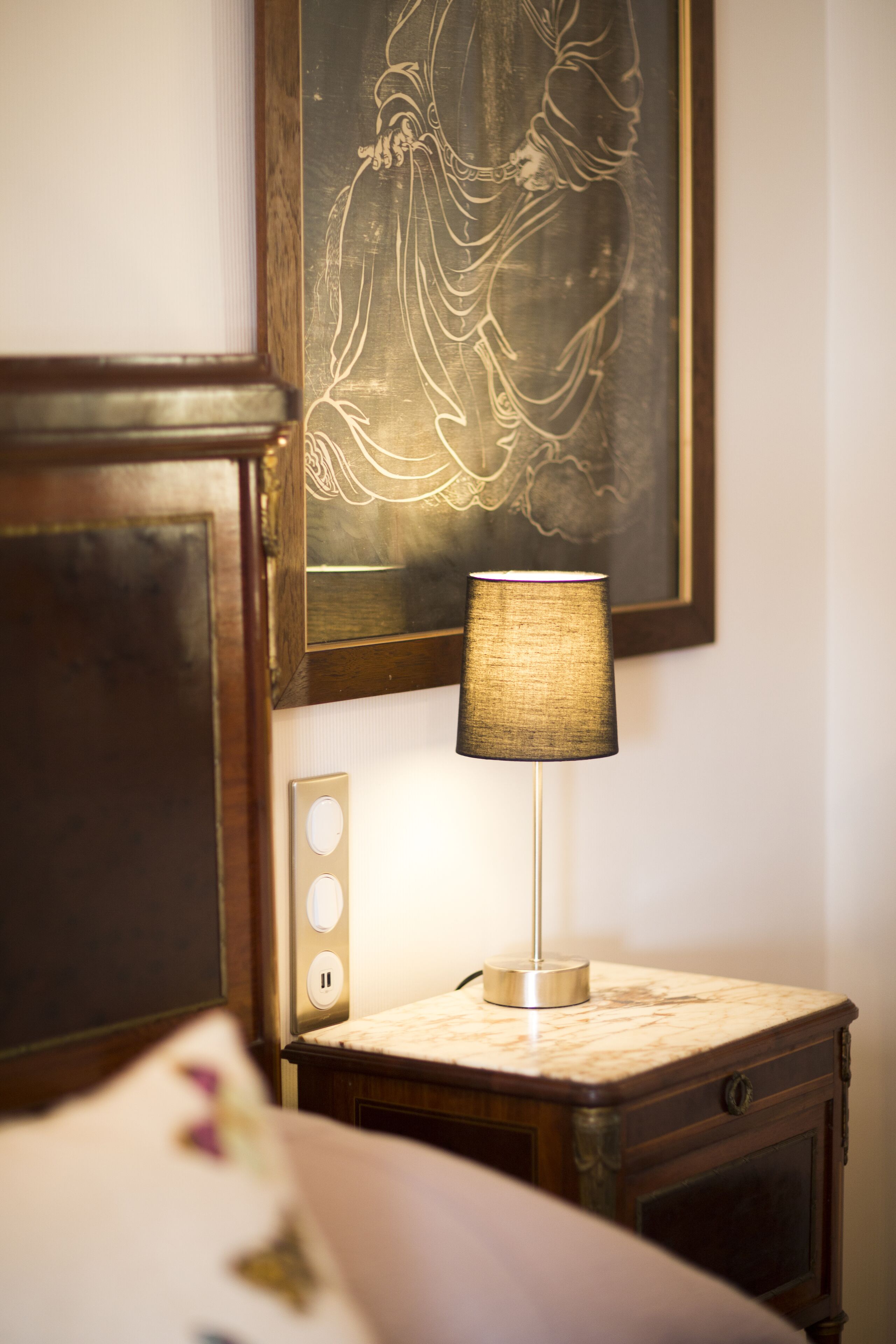 Een nachtkastje met een lamp, een stopcontact en een lijst met kunst aan de muur.