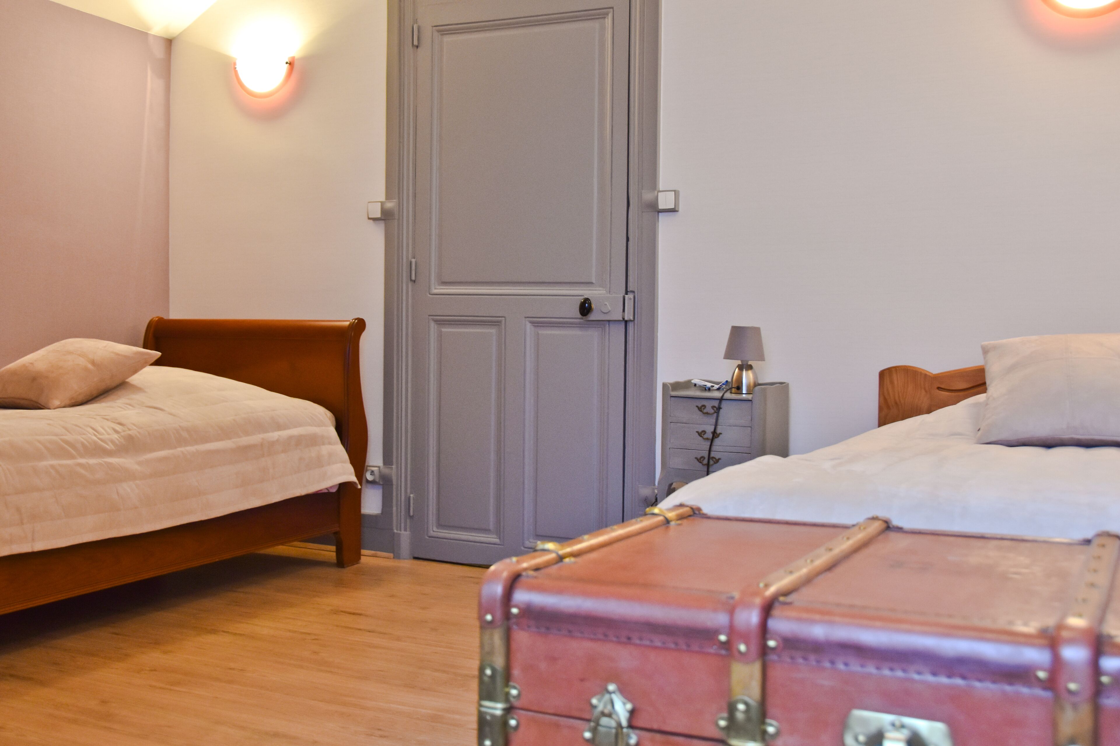 Een slaapkamer met twee bedden, een houten vloer, een grijze deur en een klein nachtkastje met een lamp.