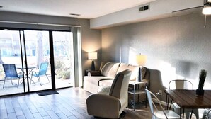 Ein modernes Wohnzimmer mit Sofa, Sesseln, Couchtisch und Esstisch. Eine Glastür führt auf eine Außenbereich mit Sitzgelegenheiten.