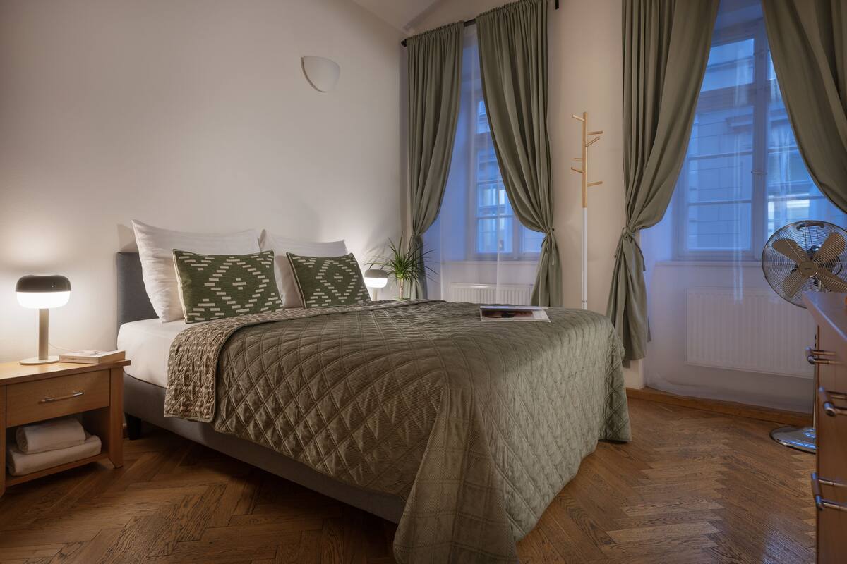 Un dormitorio con cama, mesitas de noche, una ventana con cortinas y un ventilador de pie.