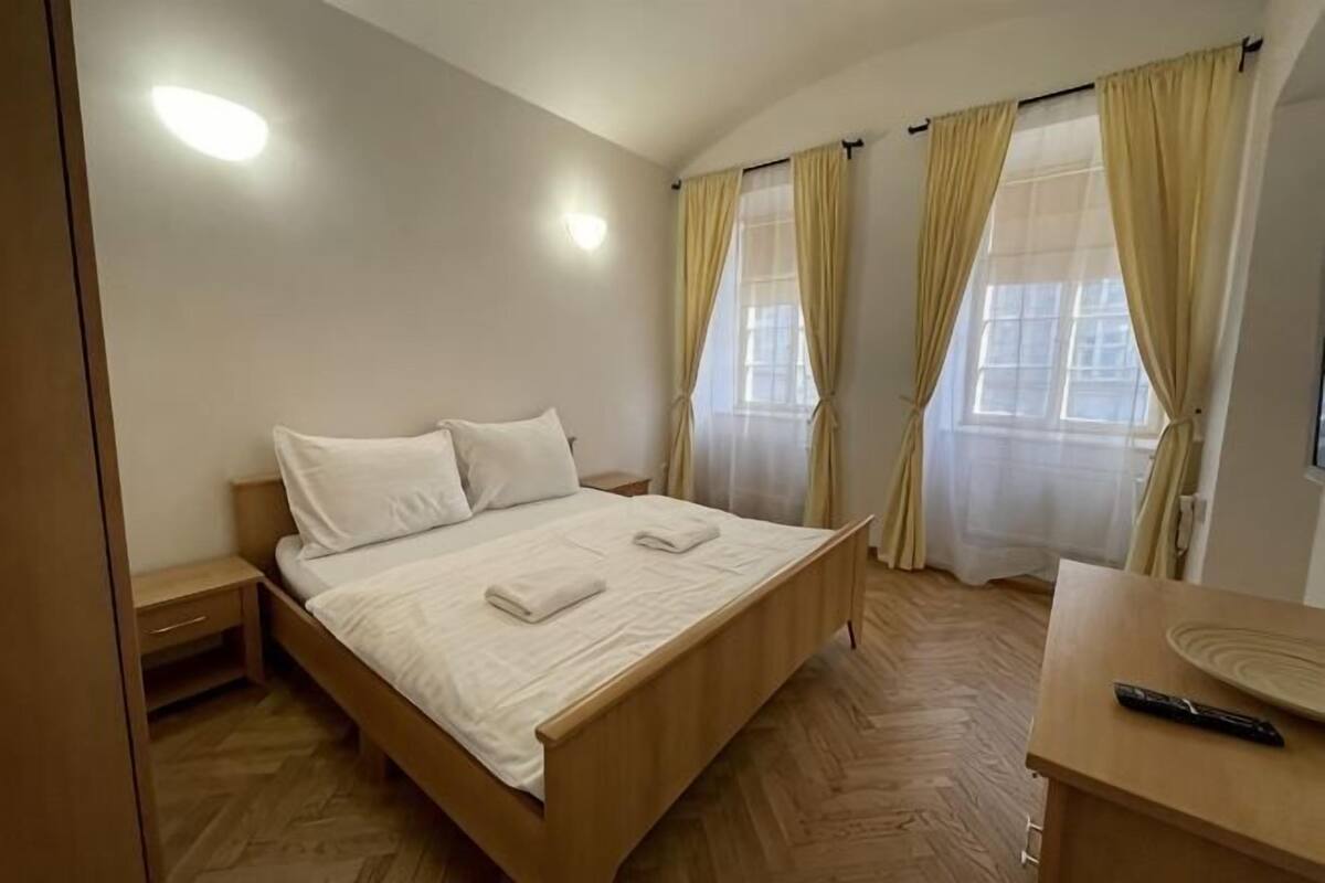 Un dormitorio con cama de madera, mesitas de noche, un escritorio con control remoto y cortinas en las ventanas.