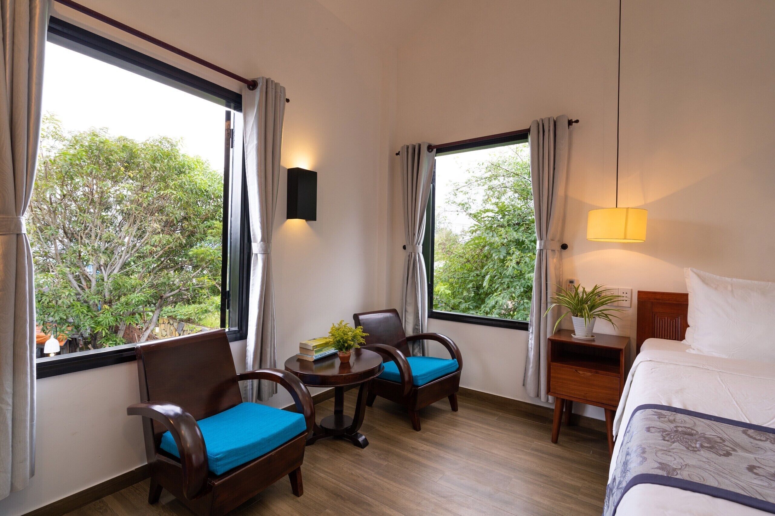 A hotel room with a bed, two chairs with blue cushions, a small table with a plant, and a nightstand with a lamp.