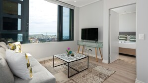 En moderne stue med stort vindue, glasbord, fladskærms-tv og seng med mønstrede puder.